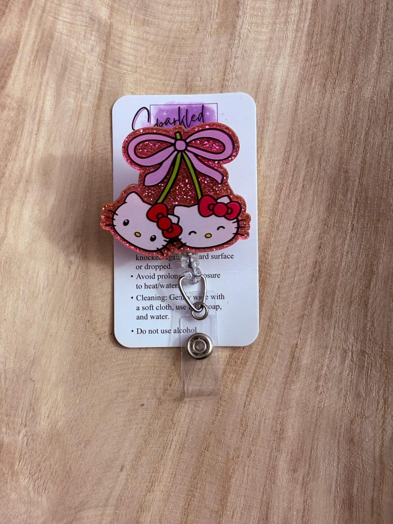 May include: Pink glitter Hello Kitty charm with a pink bow and cherries. The charm is attached to a clear plastic badge holder with a metal clip. The card behind the charm has instructions for care: "knocked against a hard surface or dropped. Avoid prolonged exposure to heat/water. Cleaning: Gently wipe with a soft cloth, use soap, and water. Do not use alcohol."