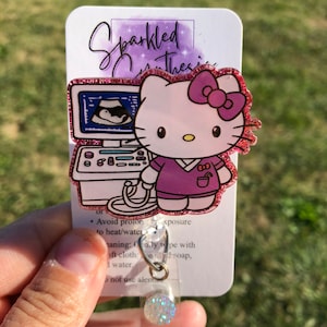 May include: A pink glitter Hello Kitty badge reel with a purple shirt and a pink bow. The Hello Kitty is holding a small ultrasound machine with a baby inside. The badge reel has a clear plastic cover and a sparkly silver charm.