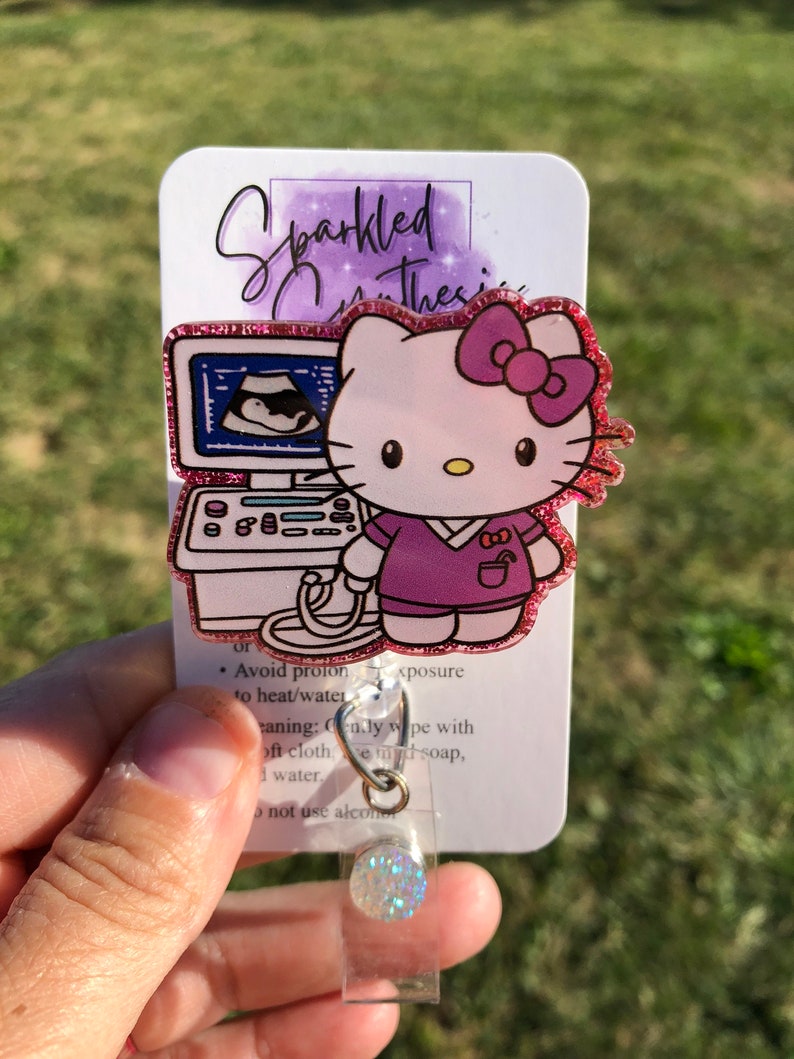 May include: A pink glitter Hello Kitty badge reel with a purple shirt and a pink bow. The Hello Kitty is holding a small ultrasound machine with a baby inside. The badge reel has a clear plastic cover and a sparkly silver charm.