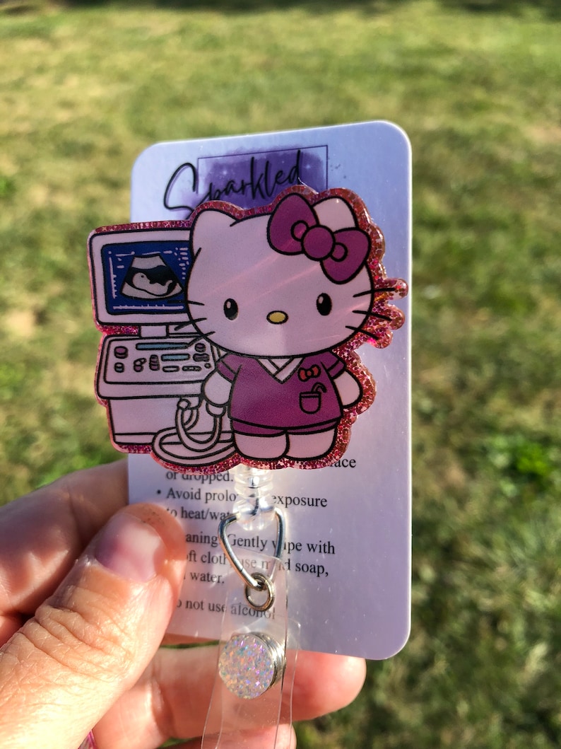 May include: A pink Hello Kitty character wearing a purple scrub top and holding a pink ultrasound machine. The character is on a white card with the word "Sparkled" in pink glitter lettering. The card also includes instructions for cleaning the badge holder. The badge holder is a clear plastic retractable reel with a sparkly white charm.