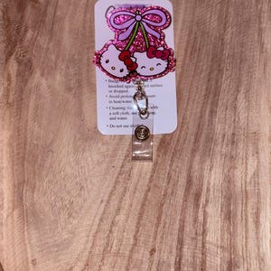 May include: Pink Hello Kitty glitter badge reel with a pink bow and two cherries. The reel has a clear plastic clip and a white card with care instructions. "Badge Reel" is written at the top of the card. The card also includes instructions to avoid prolonged exposure to heat/water, to clean with a soft cloth and water, and to not use alcohol.