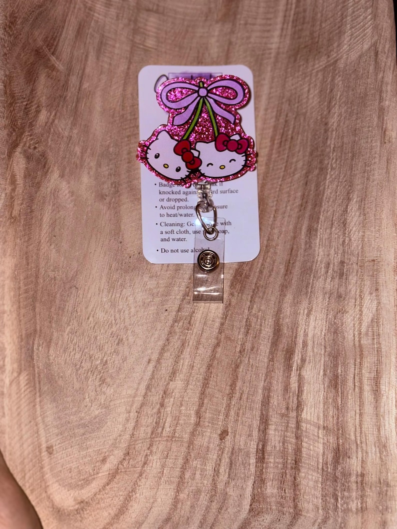 May include: Pink Hello Kitty glitter badge reel with a pink bow and two cherries. The reel has a clear plastic clip and a white card with care instructions. "Badge Reel" is written at the top of the card. The card also includes instructions to avoid prolonged exposure to heat/water, to clean with a soft cloth and water, and to not use alcohol.