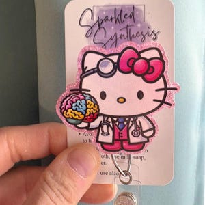 Puede incluir: Un portatarjetas con un diseño de Hello Kitty como doctora, sosteniendo una ilustración de cerebro colorido. El portatarjetas tiene el texto "Sparkled Synthesis" e incluye un soporte de plástico transparente y un botón brillante.