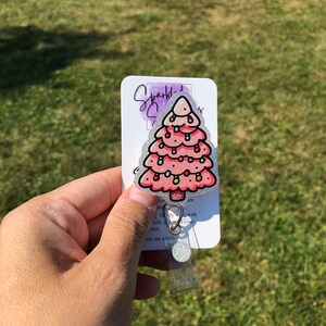 May include: A pink glitter Christmas tree with white lights and a silver glitter background. The tree is attached to a clear plastic badge holder with a silver clip. The text on the card reads "Sparkly", "Jon are ate", "Gently wipe with cloth and soap", "water", "not use alcohol".