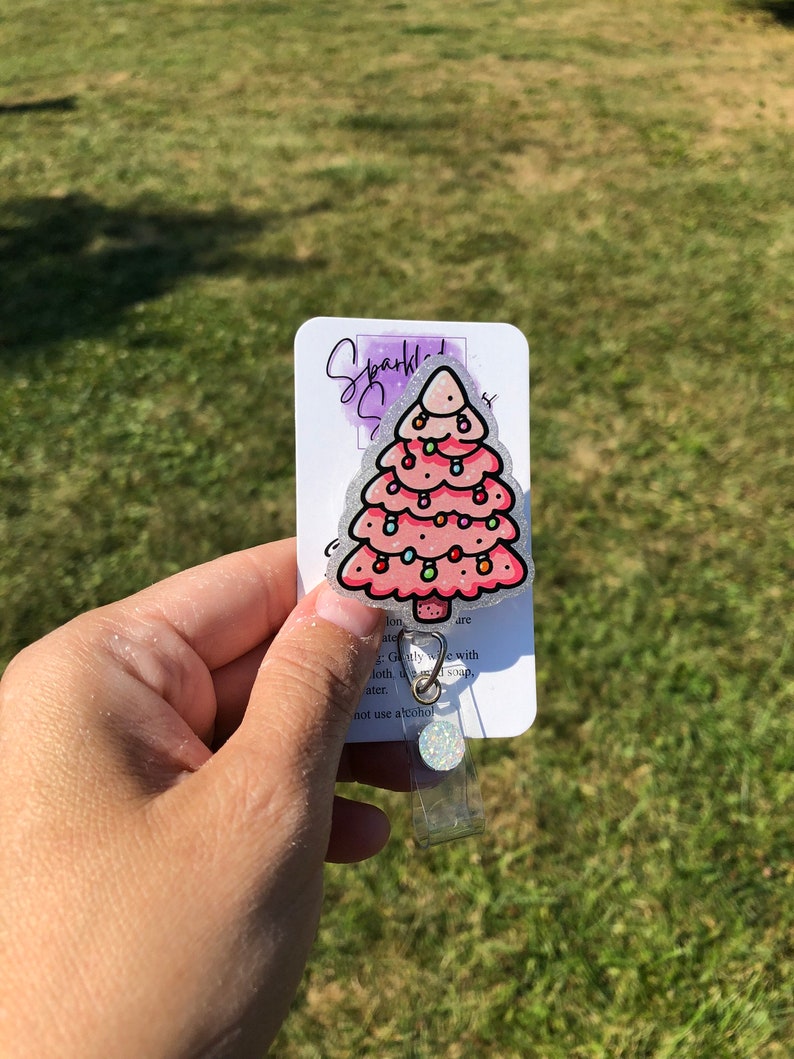 May include: A pink glitter Christmas tree with white lights and a silver glitter background. The tree is attached to a clear plastic badge holder with a silver clip. The text on the card reads "Sparkly", "Jon are ate", "Gently wipe with cloth and soap", "water", "not use alcohol".
