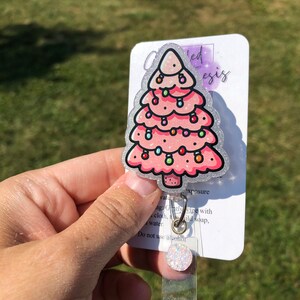 May include: A pink glitter Christmas tree with colorful lights, a silver glitter background, and the text "watu ed esis" and "xposure" on a white card. The card also includes instructions for cleaning the badge holder: "Gently wipe with cloth, mild soap, water. Do not use alcohol."