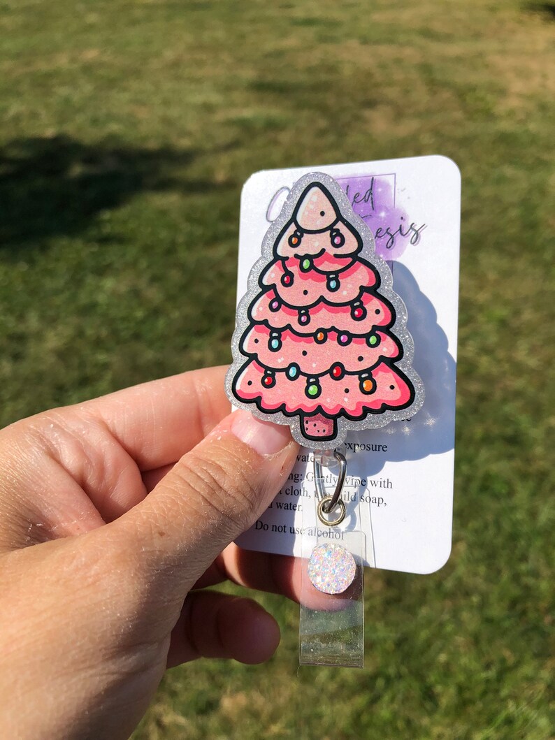 May include: A pink glitter Christmas tree with colorful lights, a silver glitter background, and the text "watu ed esis" and "xposure" on a white card. The card also includes instructions for cleaning the badge holder: "Gently wipe with cloth, mild soap, water. Do not use alcohol."