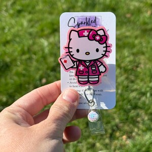 Op de afbeelding: Roze Hello Kitty verpleegster badge reel met een wit kruis en een wit medisch dossier. De badge reel heeft een transparante plastic hoes en een glinsterende, regenboogkleurige knop.