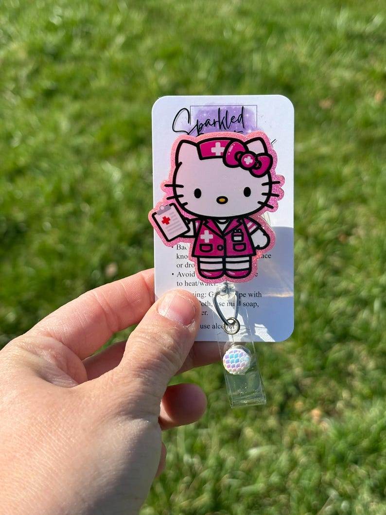 Op de afbeelding: Roze Hello Kitty verpleegster badge reel met een wit kruis en een wit medisch dossier. De badge reel heeft een transparante plastic hoes en een glinsterende, regenboogkleurige knop.