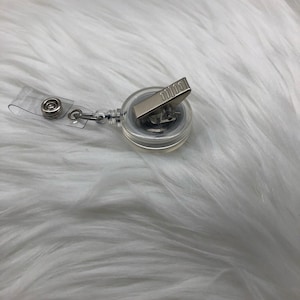 May include: A clear retractable badge holder with a silver clip. The holder is on a white fuzzy surface.