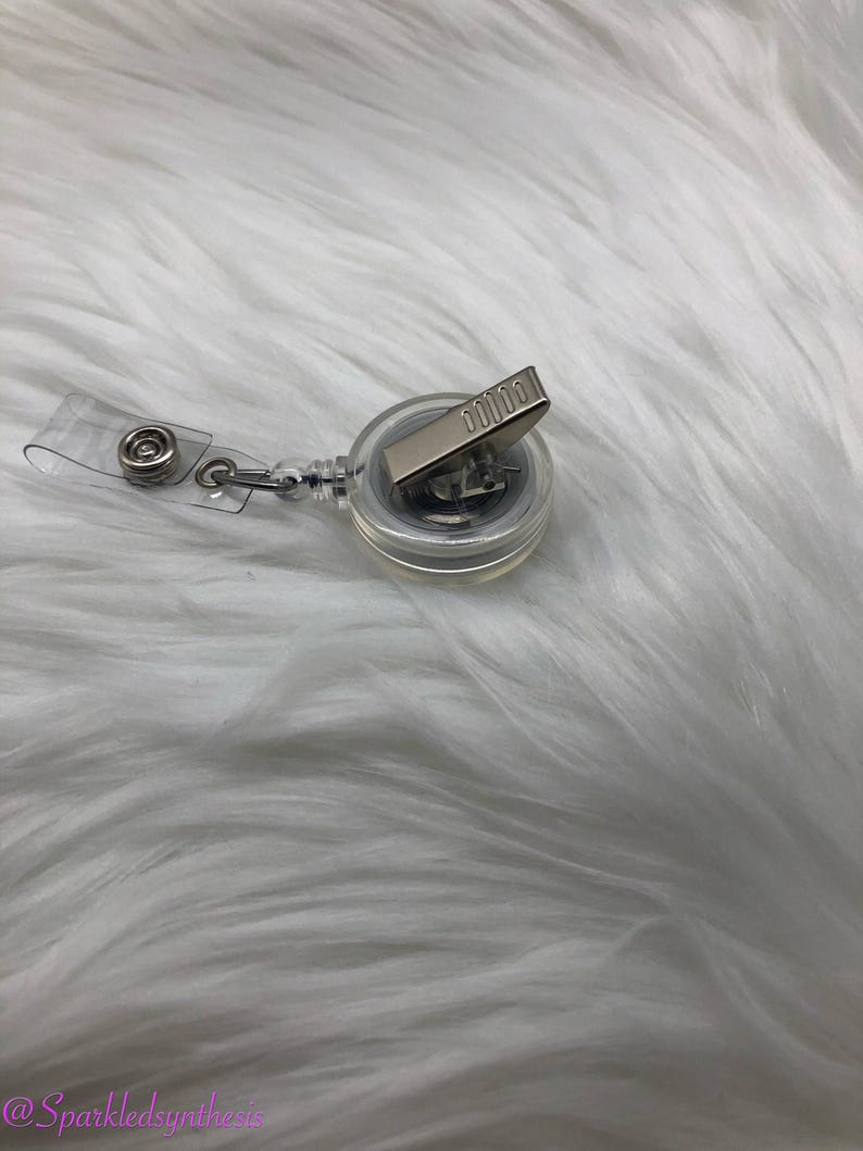 May include: A clear retractable badge holder with a silver clip. The holder is on a white fuzzy surface.