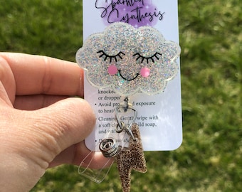 Può includere: Una nuvola bianca scintillante con un viso sorridente e guance rosa. La nuvola è attaccata a un ciondolo a forma di fulmine dorato glitterato e a un porta badge in plastica trasparente. Il testo "Sparkled Synthesis" è stampato su una carta bianca dietro la nuvola.