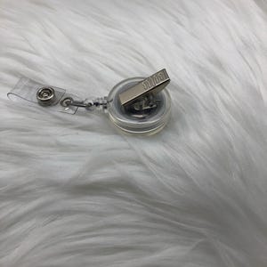 Op de afbeelding: Een transparante intrekbare badgehouder met een zilveren clip. De houder bevindt zich op een wit, pluche oppervlak.