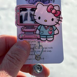 May include: A pink Hello Kitty character wearing a blue scrub top and a pink bow. The character is standing in front of a pink and white medical imaging machine. The image is on a white card with the text "Sparkled Synthesis" at the top. The card is attached to a clear plastic badge holder.