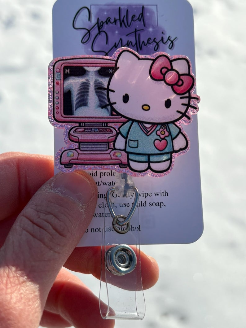 May include: A pink Hello Kitty character wearing a blue scrub top and a pink bow. The character is standing in front of a pink and white medical imaging machine. The image is on a white card with the text "Sparkled Synthesis" at the top. The card is attached to a clear plastic badge holder.