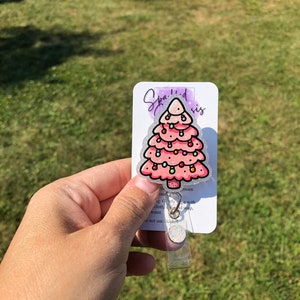 May include: A pink glitter Christmas tree with colorful lights and ornaments. The tree is on a white card with the text "Sparkled sis" and instructions for care. The card is attached to a clear plastic badge holder.