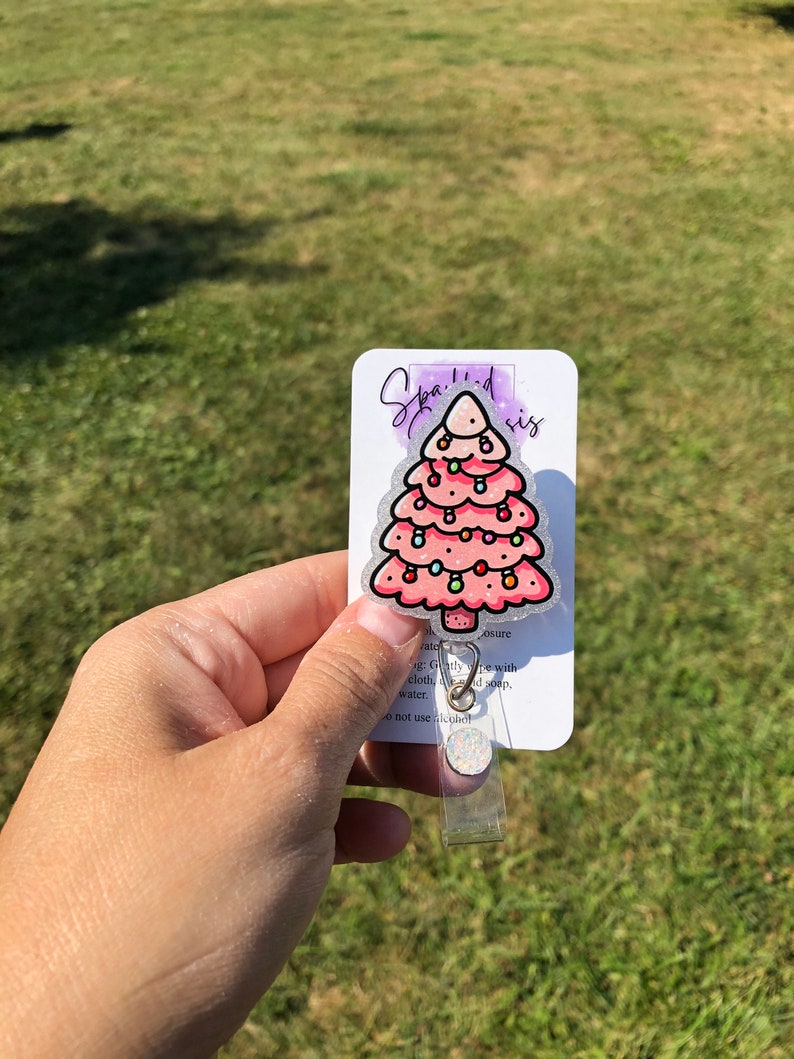 May include: A pink glitter Christmas tree with colorful lights and ornaments. The tree is on a white card with the text "Sparkled sis" and instructions for care. The card is attached to a clear plastic badge holder.