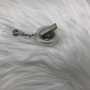 May include: A clear retractable badge holder with a silver clip. The holder is on a white fuzzy surface.