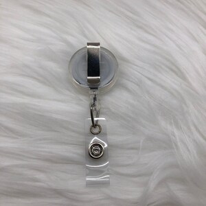 Op de afbeelding: Een transparante intrekbare badgehouder met een zilveren clip en een zilveren cirkel met een ontwerp in het midden.