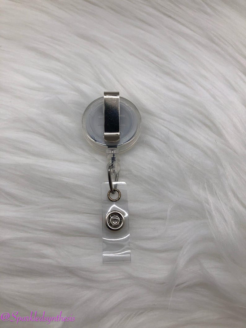 Op de afbeelding: Een transparante intrekbare badgehouder met een zilveren clip en een zilveren cirkel met een ontwerp in het midden.