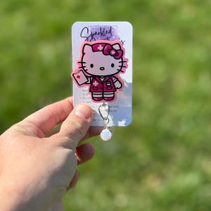 Op de afbeelding: Roze Hello Kitty verpleegster badge reel met een glinsterende afwerking. De reel heeft een Hello Kitty-personage dat is gekleed als verpleegster met een rood kruis op haar hoed en een klembord in haar hand. De tekst "Sparkled" is zichtbaar op de kaart achter de reel.