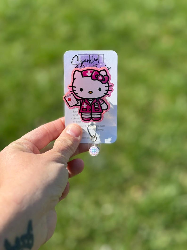 Op de afbeelding: Roze Hello Kitty verpleegster badge reel met een glinsterende afwerking. De reel heeft een Hello Kitty-personage dat is gekleed als verpleegster met een rood kruis op haar hoed en een klembord in haar hand. De tekst "Sparkled" is zichtbaar op de kaart achter de reel.