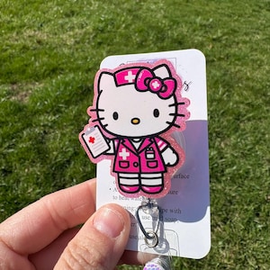 Op de afbeelding: Roze Hello Kitty verpleegster badge reel met glitter en een wit medisch kruis. De reel heeft een transparante plastic behuizing en een regenboogkleurige, iriserende charme.