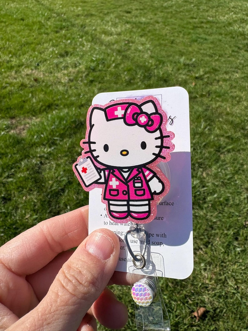 Op de afbeelding: Roze Hello Kitty verpleegster badge reel met glitter en een wit medisch kruis. De reel heeft een transparante plastic behuizing en een regenboogkleurige, iriserende charme.