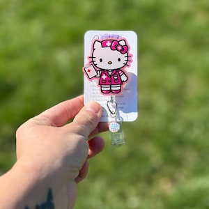 Op de afbeelding: Roze Hello Kitty verpleegster badge reel met een witte achtergrond. De reel heeft een transparante plastic behuizing met een roze en blauwe gepatenteerde knop.