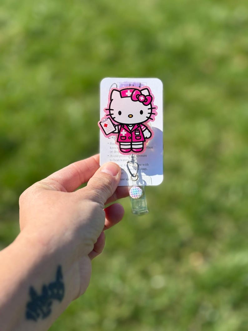 Op de afbeelding: Roze Hello Kitty verpleegster badge reel met een witte achtergrond. De reel heeft een transparante plastic behuizing met een roze en blauwe gepatenteerde knop.