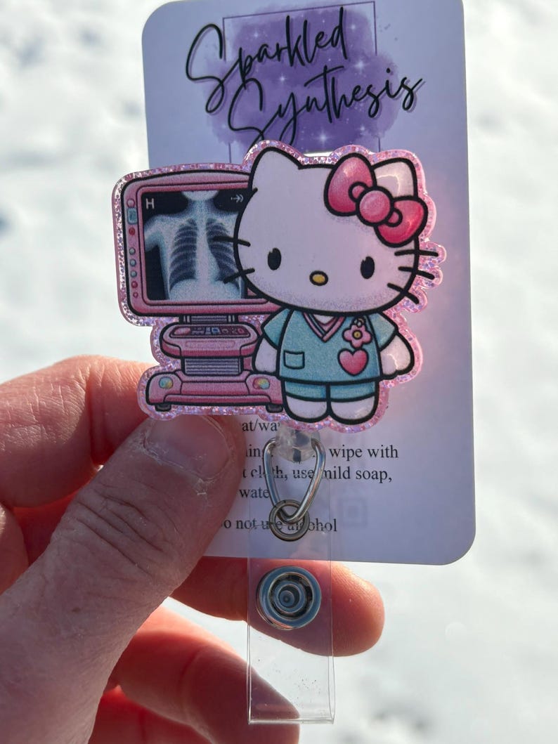 May include: Pink Hello Kitty character wearing a blue scrub top and a pink bow. She is standing in front of a pink medical monitor with an x-ray image on the screen. The image is on a clear plastic badge holder with a silver clip.