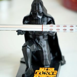 May include: Black Darth Vader figurine holding a silver pen with a red stripe and a gray grip. The figurine is kneeling and has a black base with the words "May the Force Be With You" in gold lettering.