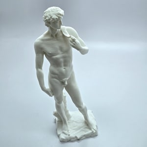 May include: A white sculpture of a nude male figure, standing on a textured base. The figure is holding a cloth over his shoulder. The statue has detailed musculature and is set against a plain background.