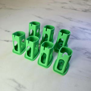 May include: Seven bright green, plastic, rectangular holders with rounded corners are arranged on a white marble surface. Each holder has a square opening at the top and a circular hole at the bottom. The sides have cut-out sections.