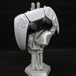 May include: A 3D printed robotic hand holding a white PlayStation 5 controller. The hand is made of a silver-colored material and has a detailed design. The controller is in a vertical position and has black buttons and a black PlayStation logo.