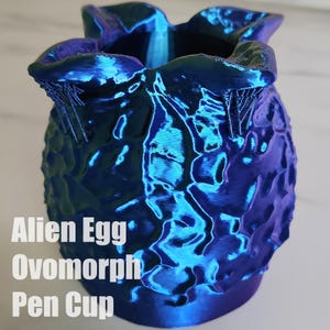 May include: A blue and purple iridescent alien egg ovomorph pen cup. The cup is shaped like an alien egg from the movie Alien.