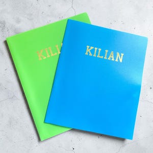 May include: Two notebooks, one bright green and one blue, are displayed on a gray surface. The word "KILIAN" is printed in gold lettering on the front of each notebook. The notebooks are slightly angled, showcasing their vibrant colors.