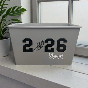 May include: A gray rectangular planter box with the numbers "2 26" in black, a winged foot graphic, and the name "Shawn" in white script. The planter is sitting on a white windowsill.