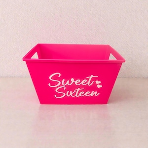 May include: A bright pink rectangular container with handles. The words "Sweet Sixteen" are printed in white script on the front, with a heart symbol. The container is empty and sits on a light surface.