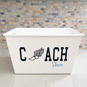 May include: A white rectangular storage bin with the word "COACH" in black block letters, a winged shoe graphic, and the name "Steve" in light blue. The bin is on a white surface with a brick wall background.