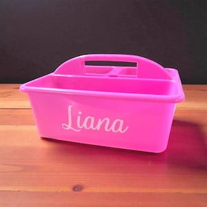 May include: A bright pink plastic storage caddy featuring a central handle and multiple interior compartments. The front of the caddy displays the name Liana printed in a white cursive script font, providing a clear view of the product's design.