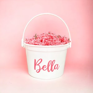 May include: A white bucket with a handle, filled with pink shredded paper. The name "Bella" is written in pink script on the front of the bucket. The background is a solid pink color.
