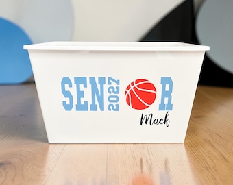 May include: White rectangular storage bin with a lid. The bin features the text "SENIOR 2027" in light blue, a red basketball graphic, and the name "Mack" in black script. The bin is on a wooden surface.