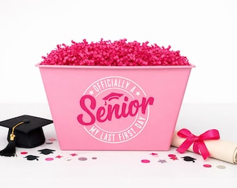 Puede incluir: Un contenedor rectangular rosa lleno de papel triturado rosa brillante. El frente presenta un gráfico circular blanco con el texto "OFFICIALLY A Senior MY LAST FIRST DAY" y un icono de birrete. Un birrete negro y un diploma enrollado están cerca.