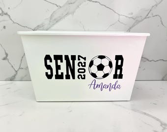 May include: A white rectangular storage bin featuring black text that reads SENIOR 2027 with a soccer ball graphic replacing the letter O. Below the graphic, the name Amanda is printed in a purple script font on the front of the container.