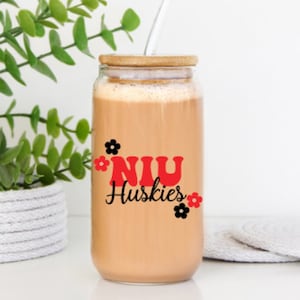 May include: A clear glass tumbler with a wooden lid and a straw, filled with a light brown beverage. The tumbler features the text "NIU Huskies" in red and black, with flower accents. The glass is sitting on a white surface.