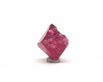 May include: A deep pink, faceted, cubic spinel crystal. The crystal is sitting on a small, gray stand.