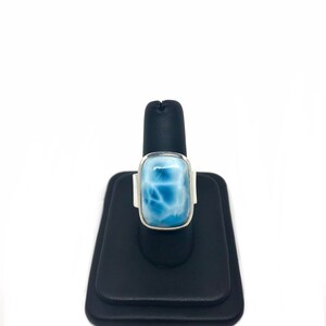 May include: A silver ring with a light blue, rectangular,  faceted gemstone. The ring is on a black ring display stand.