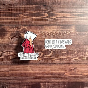 May include: Two stickers on a wooden surface. One sticker depicts a figure in a red robe and white bonnet, giving the middle finger, with the text "Nolite Te Bastardes Carborundorum." The other sticker reads "Don't Let The Bastards Grind You Down."