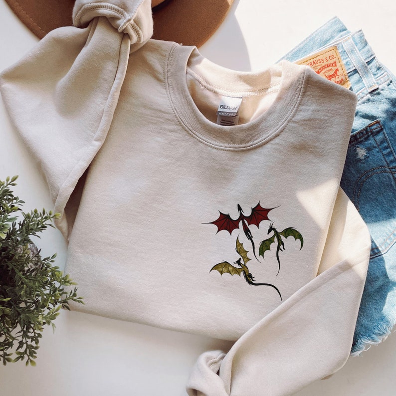 May include: A beige sweatshirt with a graphic of three dragons flying. The dragons are red, green, and yellow.